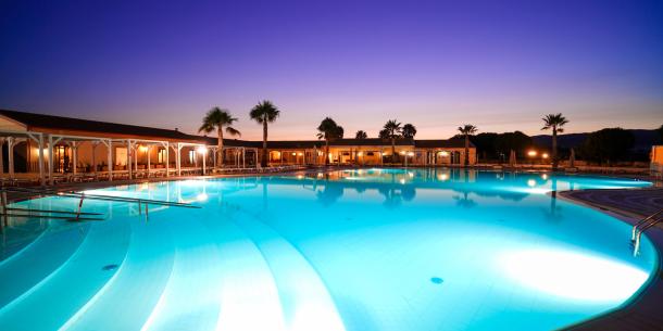 Piscina illuminata al tramonto, atmosfera serena con palme e cielo colorato.