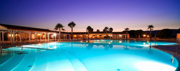 Piscina illuminata al tramonto, atmosfera serena con palme e cielo colorato.